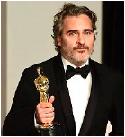 Fotografia de Joaquin Phoenix, de terno escuro, camisa branca e gravata-borboleta, sorrindo e segurando a estatueta do Oscar que ganhou como melhor ator em 2020.